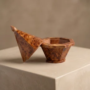 A handcrafted, faceted arar wood keepsake box by Artbeldy, resting on a minimalist beige micro-cement plinth. The geometric lid leans elegantly against the base, showcasing the rich, natural grain of the solid Moroccan wood under warm, diffused lighting.